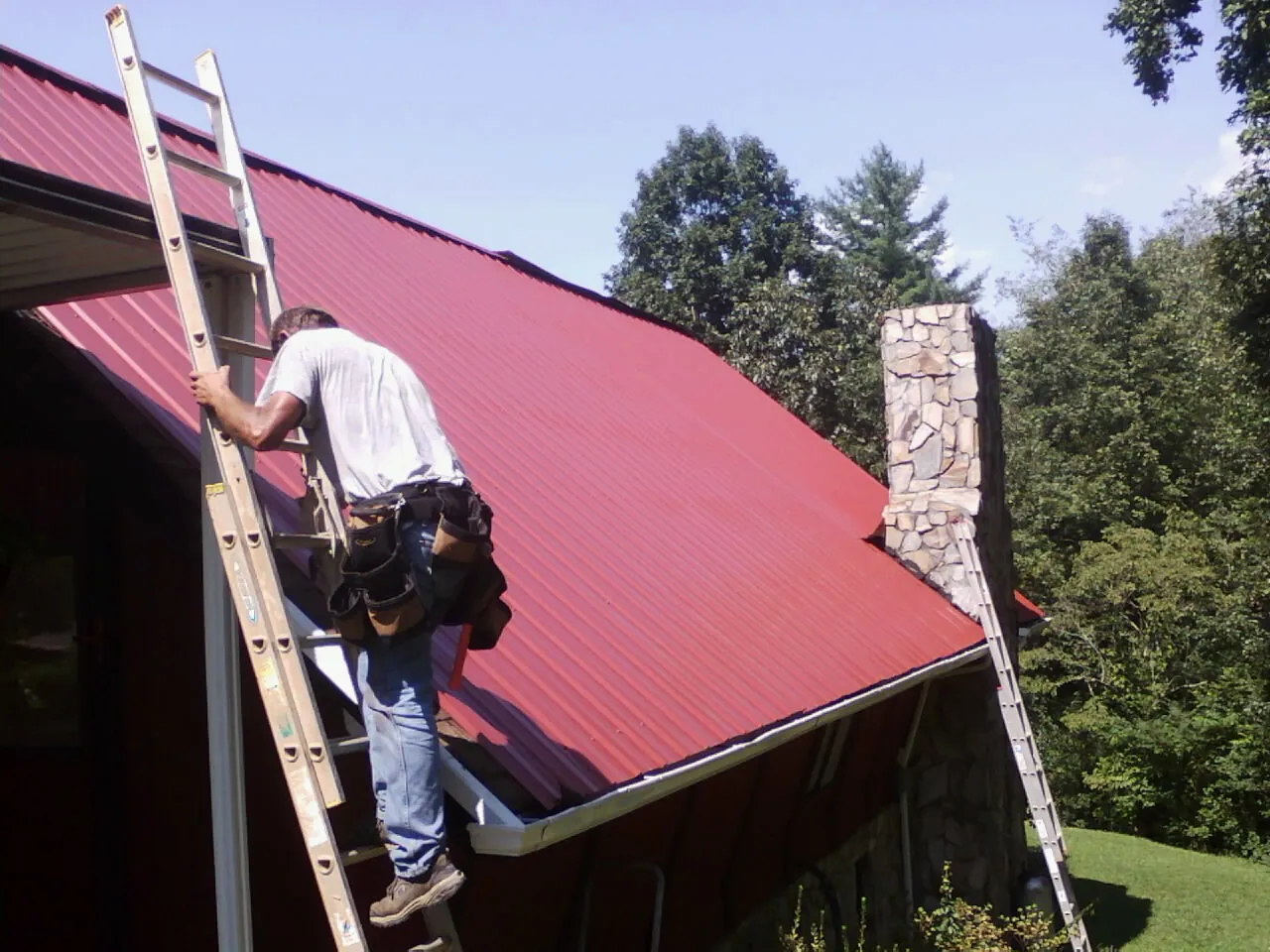 Roofer on a ladder beside a red metal roof and stone chimney — Soffit & Fascia Repair by Roofing Brothers in Pandora